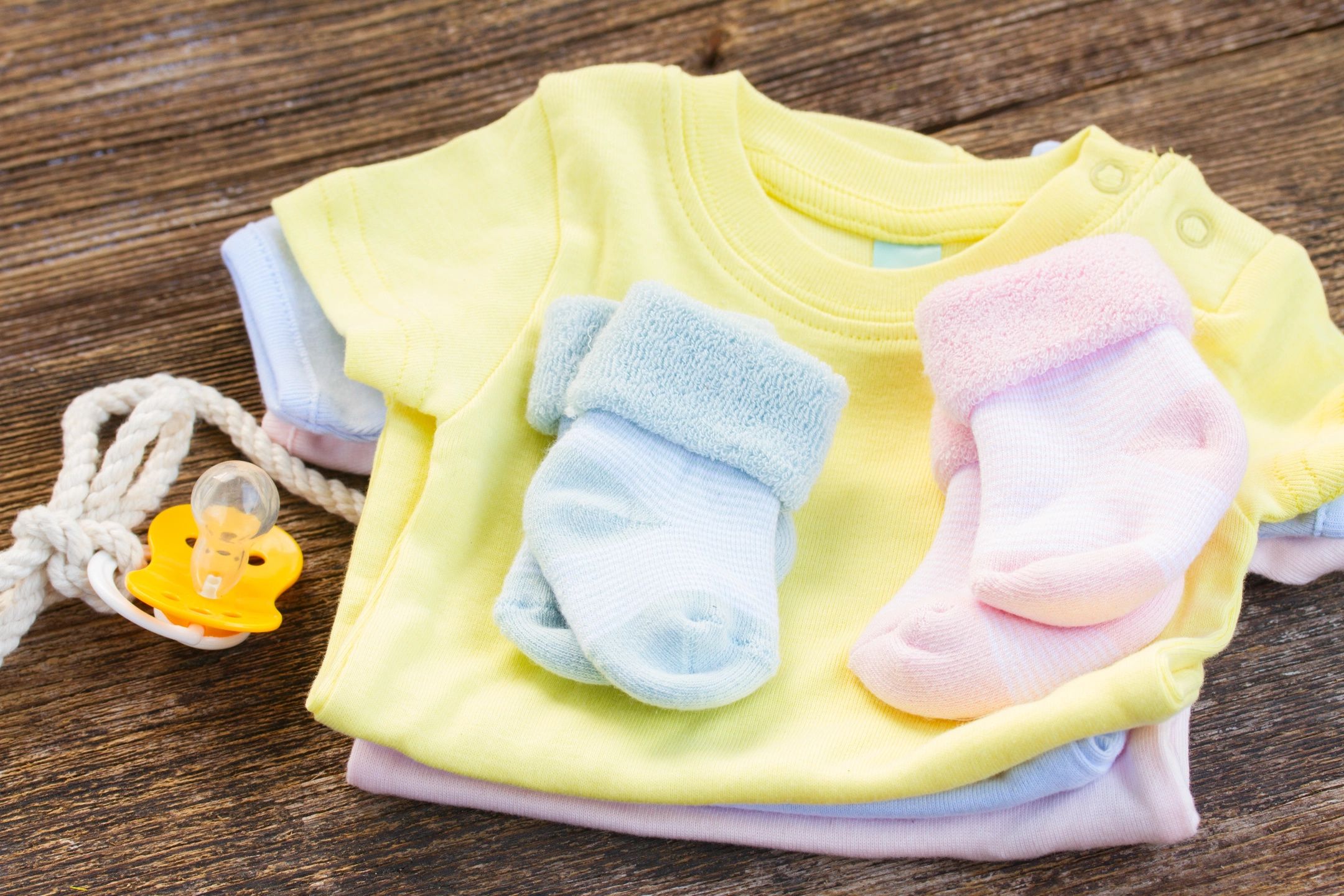 baby clothes sitting on a table. A yellow shirt, a pacifier, a blue pair of socks and a pink pair of socks.