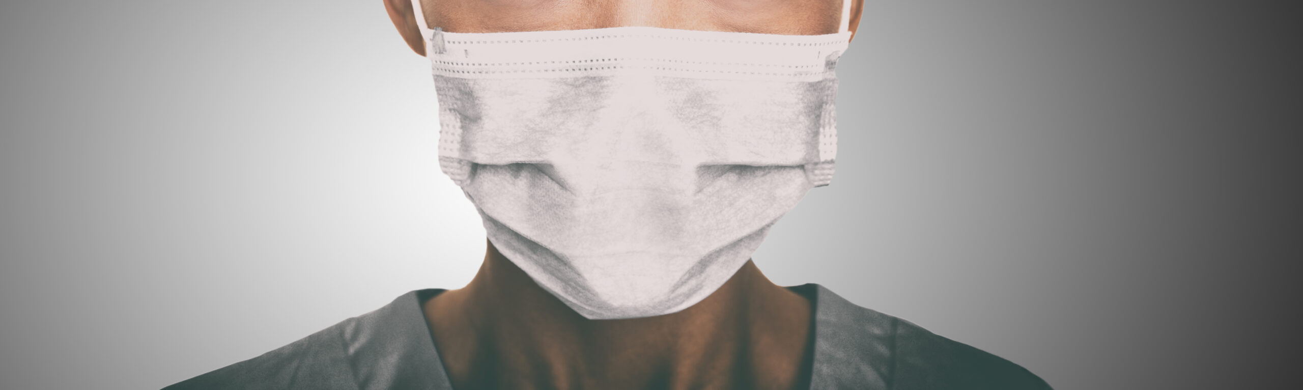 A healthcare worker wears a mask while facing the camera in front of a neutral background.
