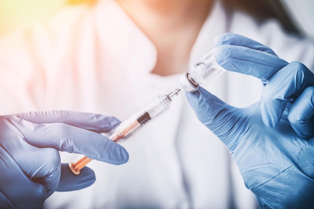 Two hands wearing surgical gloves holding syringe and drawing from a vial